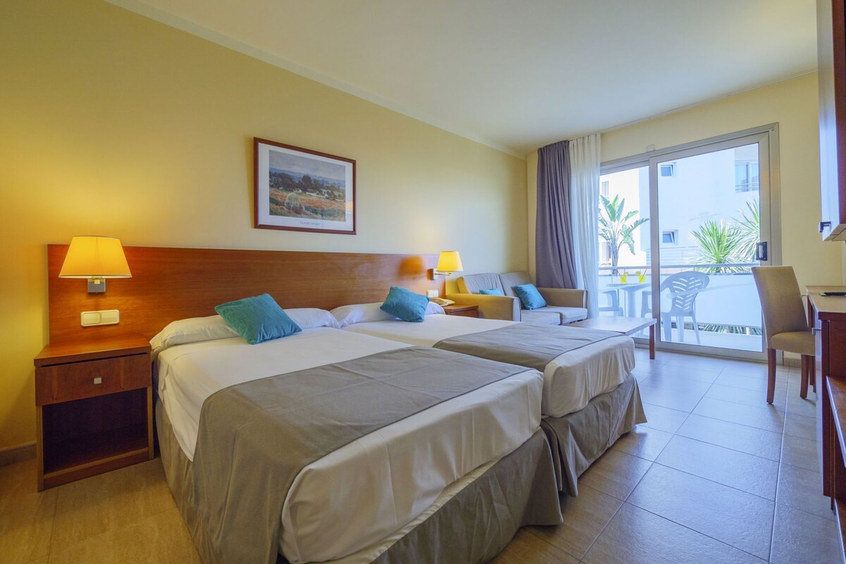 A hotel room with two beds, a desk, a chair, a balcony with a view, and a framed picture on the wall.