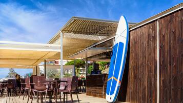 Een terras met tafels en stoelen, een surfboard tegen een houten muur en een overdekte patio.