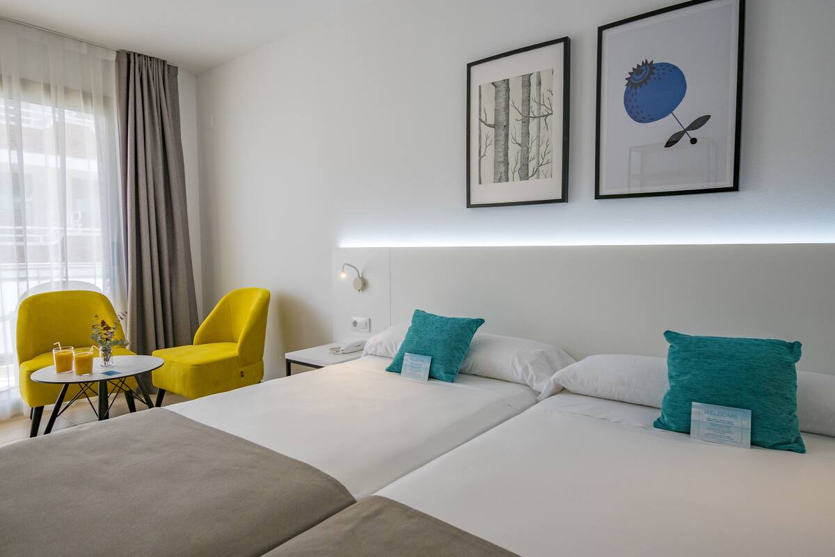 A hotel room with two beds, a small table with drinks, a chair, and artwork on the wall.
