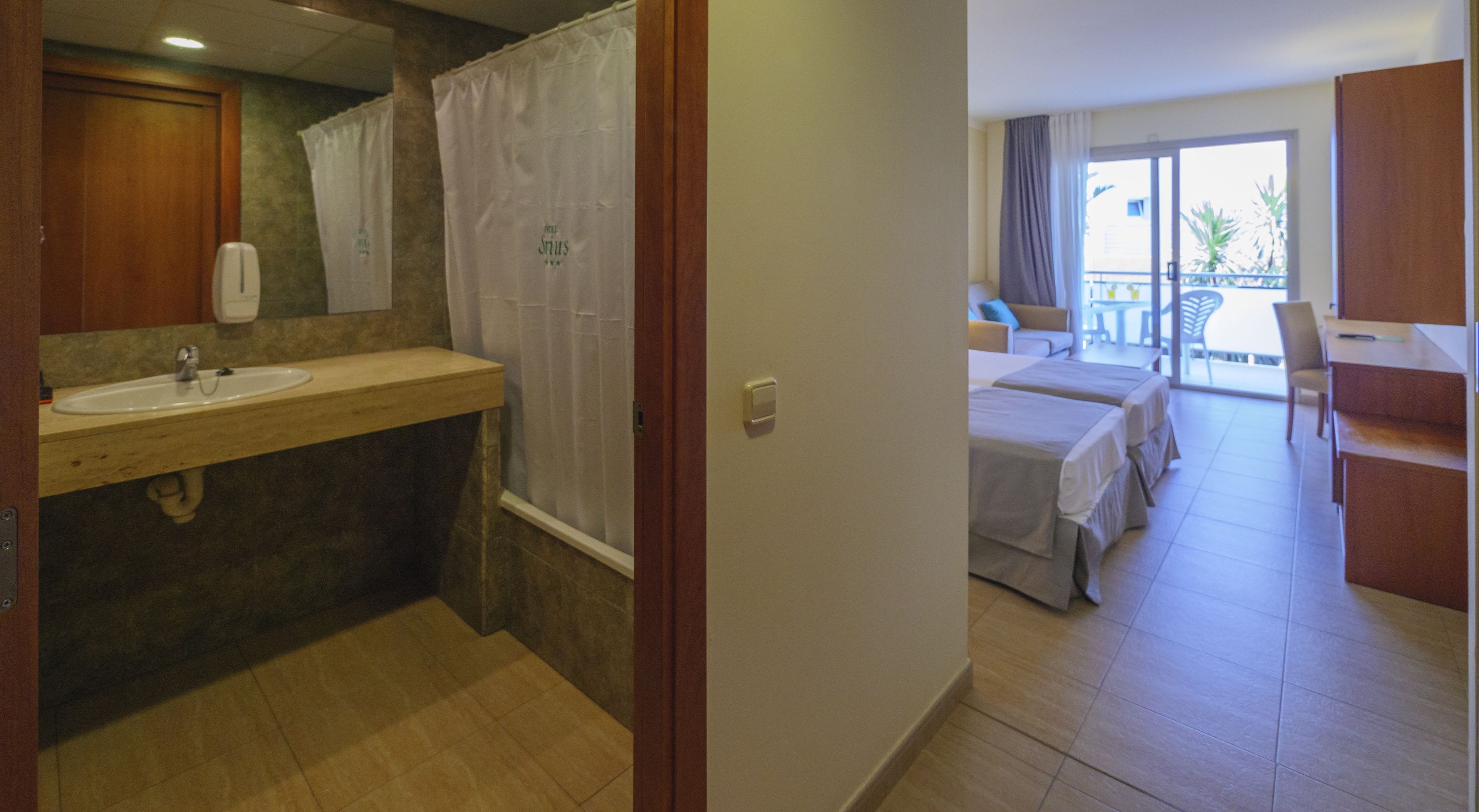 Une salle de bain équipée d’un lavabo, d’un miroir et d’un rideau de douche. Elle donne sur une chambre avec deux lits et un balcon.