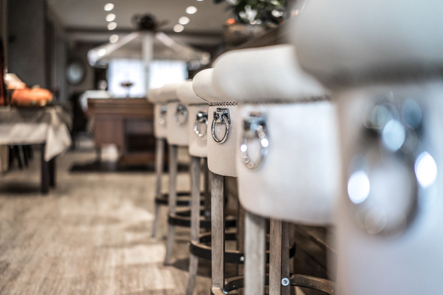 Una hilera de taburetes blancos con patas de metal y un asiento de aro, colocados en un ambiente de restaurante con suelo de madera y mesas al fondo.