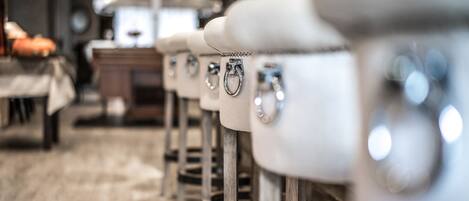 A row of white bar stools with metal legs and a ring handle, arranged in a restaurant setting with a wooden floor and tables in the background.