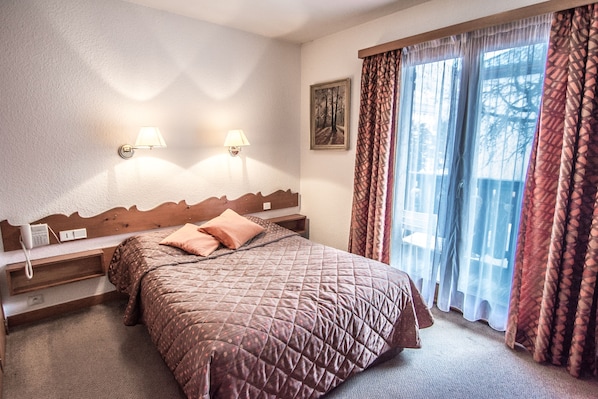 Habitación de hotel con cama, dos lámparas adosadas a la pared, teléfono, un cuadro y ventana con cortinas estampadas.
