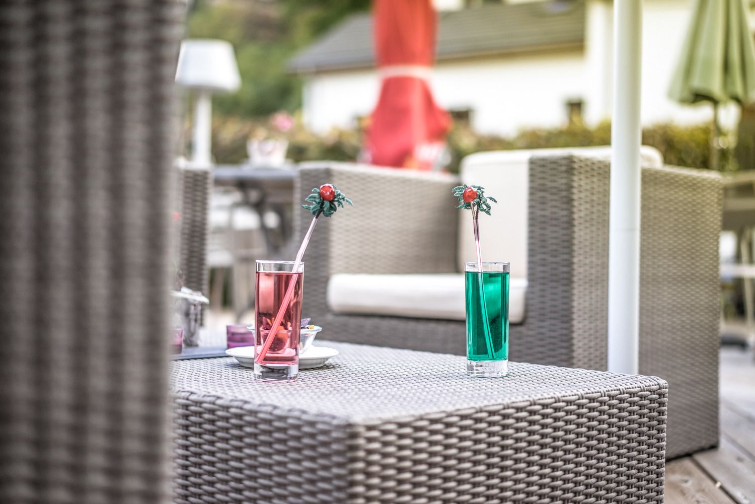Two drinks on a wicker table with a red umbrella and a white chair in the background.