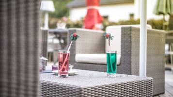 Duas bebidas sobre uma mesa de vime, com um guarda-sol vermelho e uma cadeira branca ao fundo.