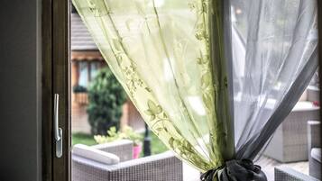A window with a floral sheer curtain tied back, revealing a patio area with wicker furniture and a garden.