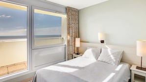 A hotel room with a large window, a bed with white linens, two bedside lamps, and a view of the beach.