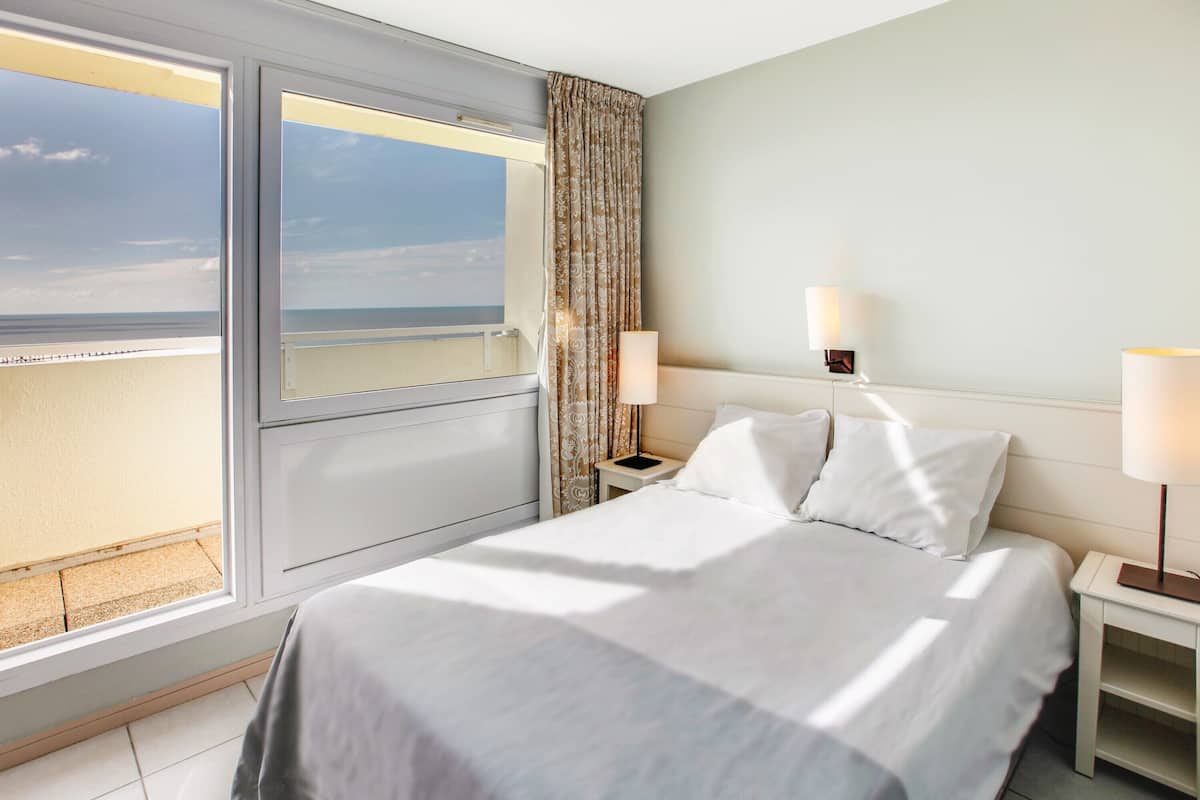 Habitación de hotel con un ventanal grande, una cama con ropa blanca, dos lámparas de noche y vistas a la playa.