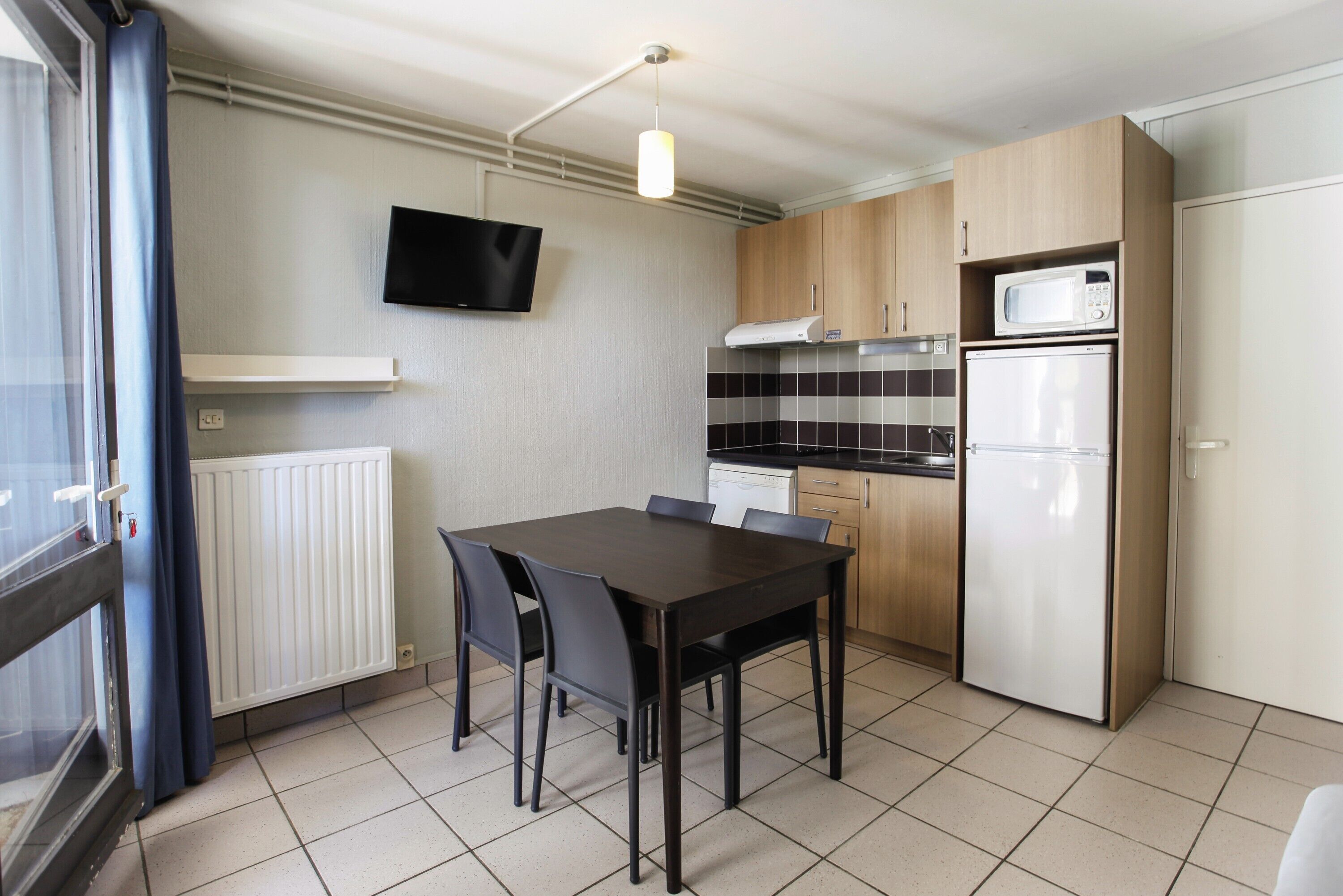 A compact kitchen with a dining table, chairs, a microwave, a refrigerator, and a TV mounted on the wall.