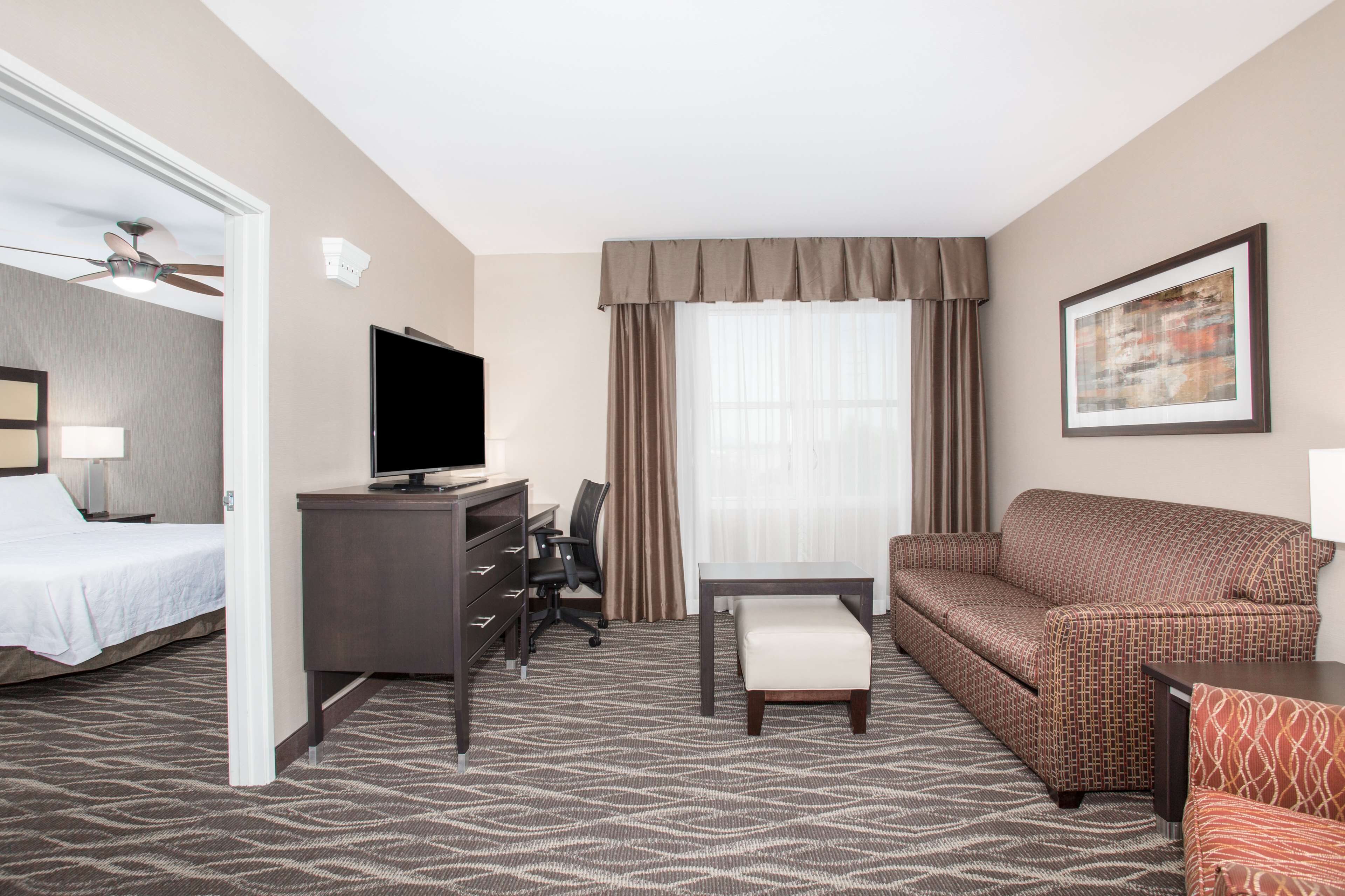 A hotel room with a bed, a desk with a TV, a sofa, a chair, and a window with curtains.