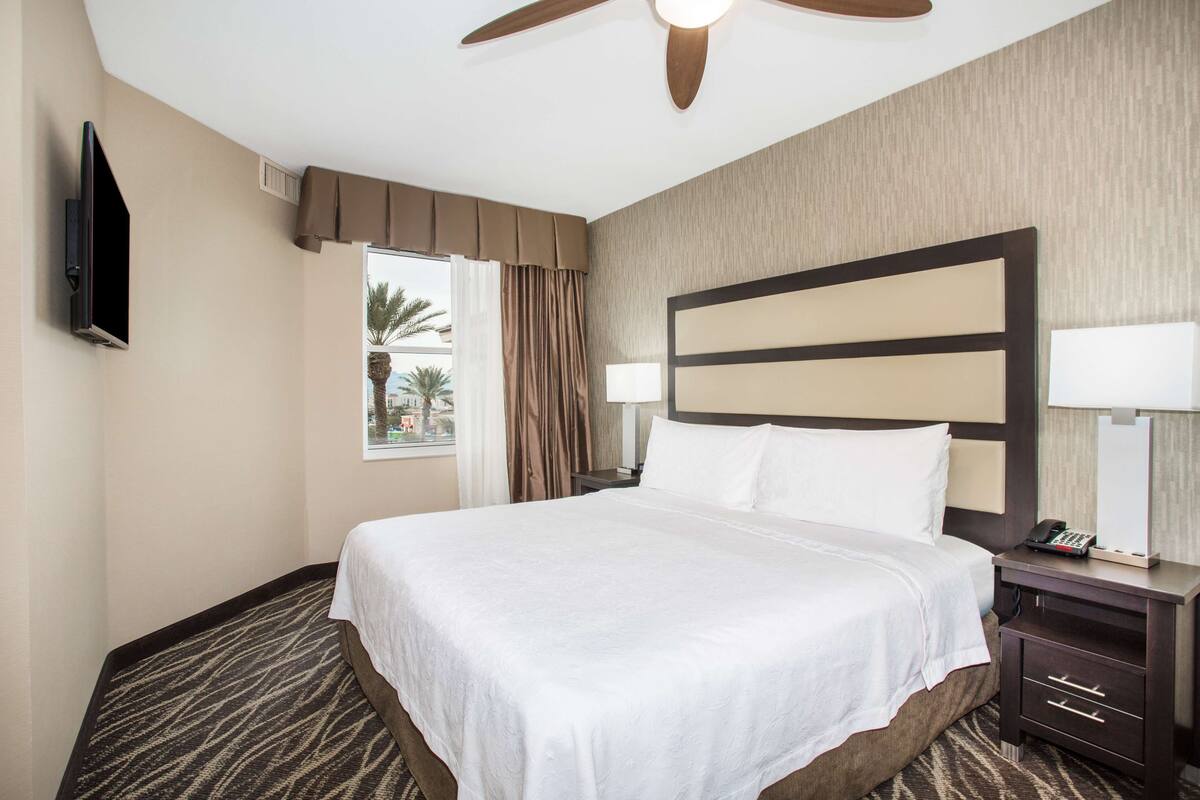 A hotel room with a large bed, a ceiling fan, a TV, a nightstand, and a window with curtains.