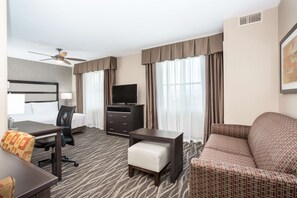 A hotel room with a bed, a desk with a chair, a sofa, a TV, and a ceiling fan. - Homewood Suites by Hilton Henderson South Las Vegas (Henderson)