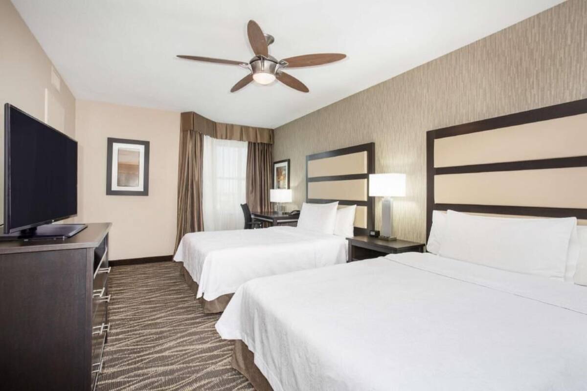 A hotel room with two beds, a desk with a TV, a ceiling fan, and a large mirror.