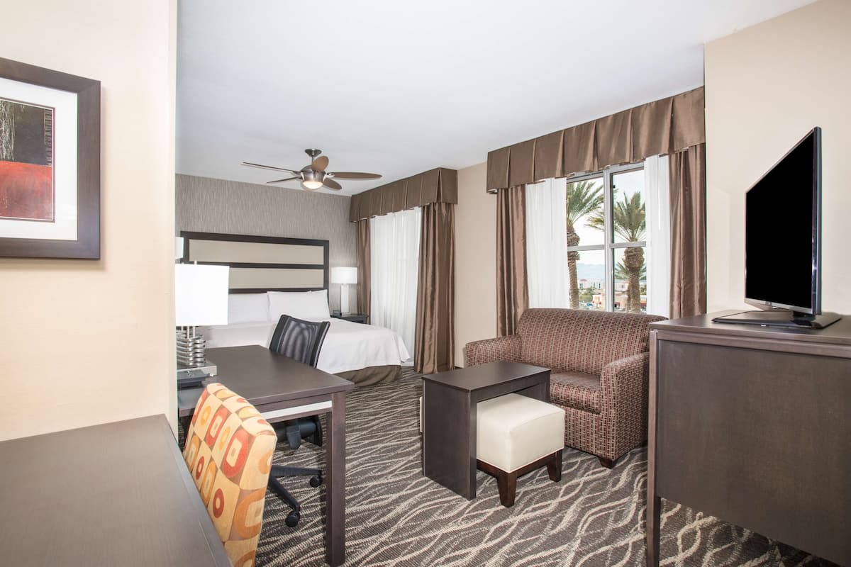 A hotel room with a bed, desk, chair, TV, and a view of palm trees.