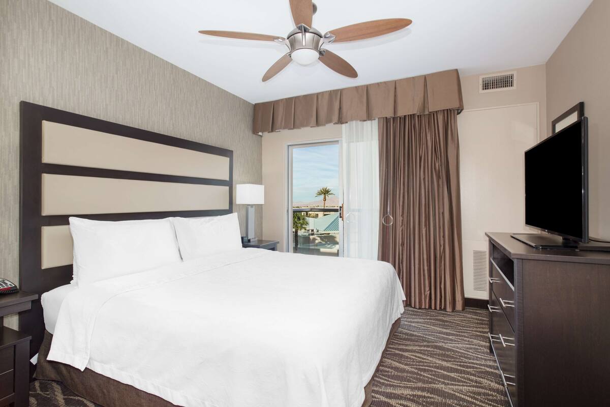 A hotel room with a large bed, a ceiling fan, a television, and a view of the outdoors through a window.