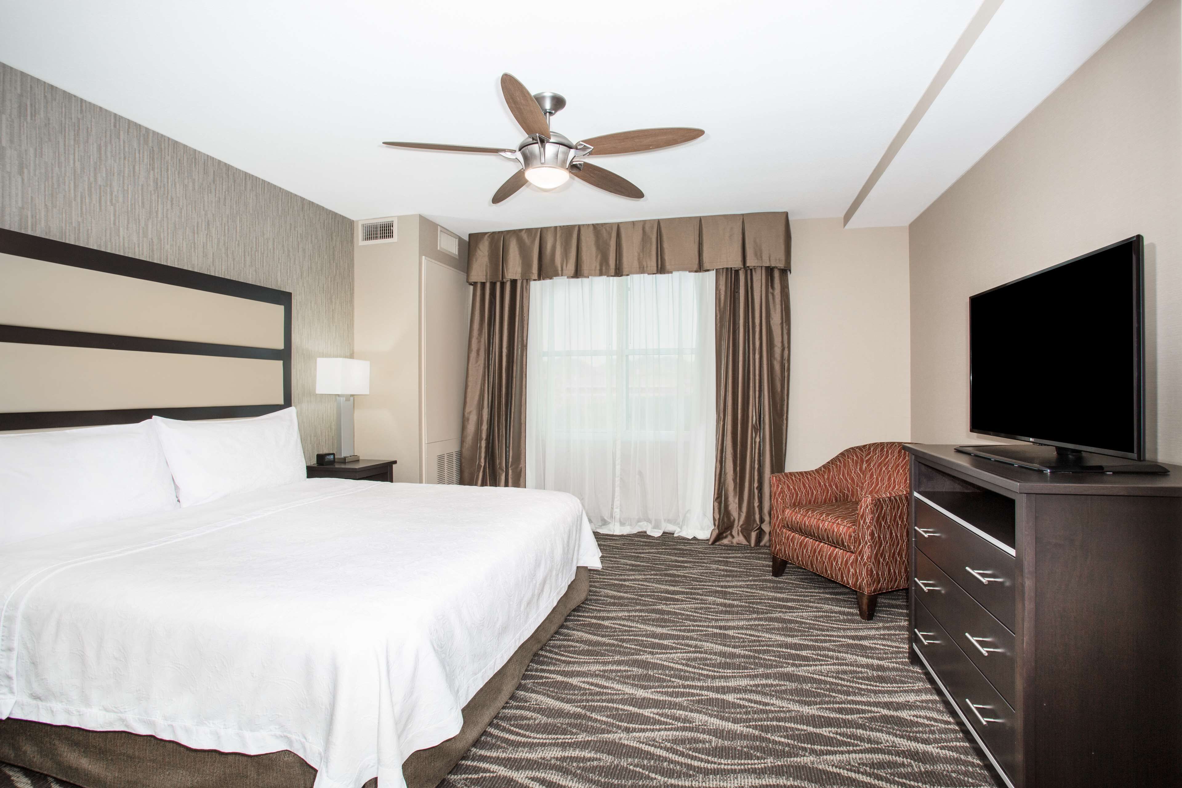 Une chambre d’hôtel avec un grand lit, un ventilateur de plafond, un téléviseur à écran plat, un tapis à motifs et un fauteuil marron.