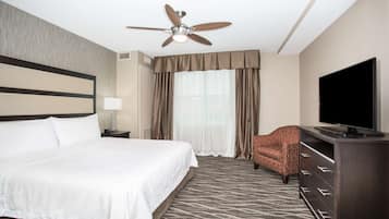 A hotel room with a large bed, a ceiling fan, a flat-screen TV, a patterned rug, and a brown armchair.