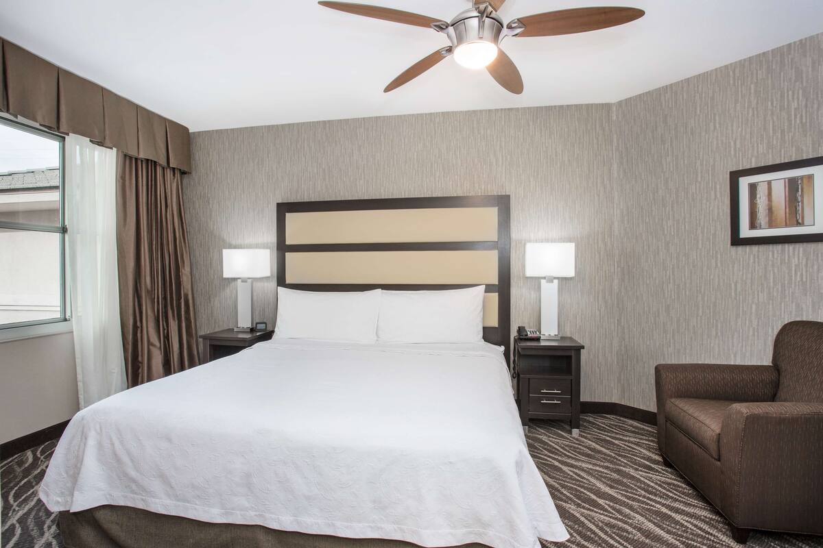 A hotel room with a large bed, two bedside tables with lamps, a ceiling fan, a brown armchair, and a framed picture on the wall.
