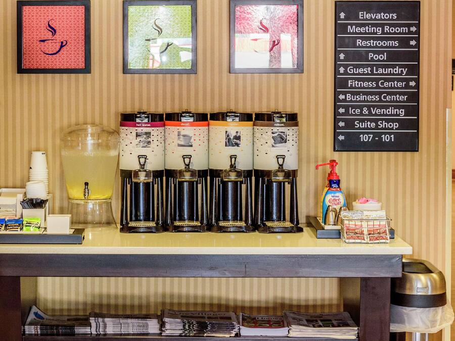 A coffee station with multiple dispensers, a large pitcher, and a coffee machine. There is a sign with directions to various hotel amenities.