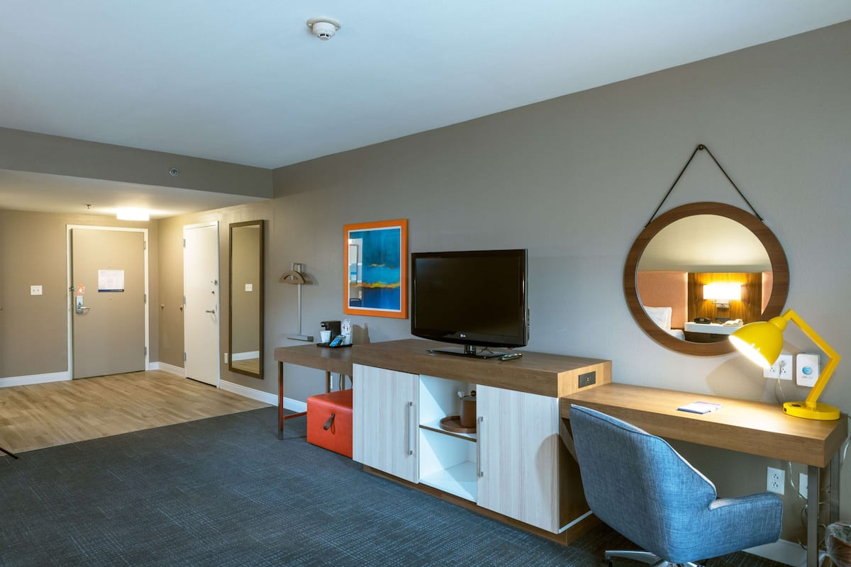 Ett hotellrum med ett skrivbord i trä, en tv, en spegel och en röd förvaringslåda.
