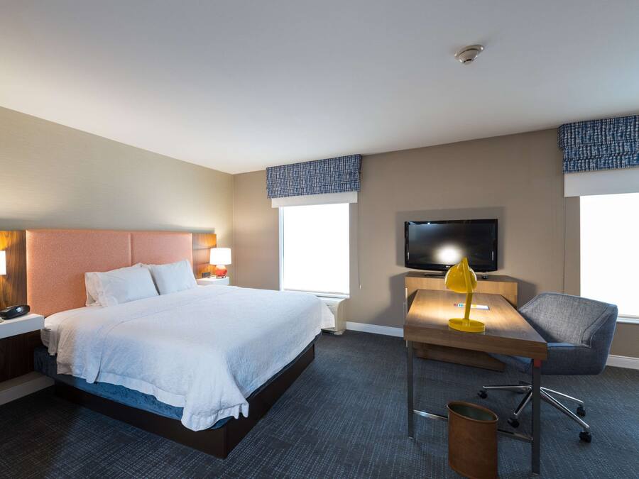Een hotelkamer met een bed, nachtkastje, lamp, televisie, bureau, stoel en een raam.