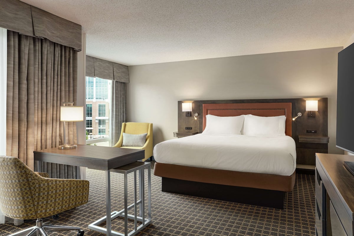 Ein Hotelzimmer mit einem großen Bett, einem Schreibtisch mit Stuhl, einem Fernseher und einem Fenster mit Vorhängen.