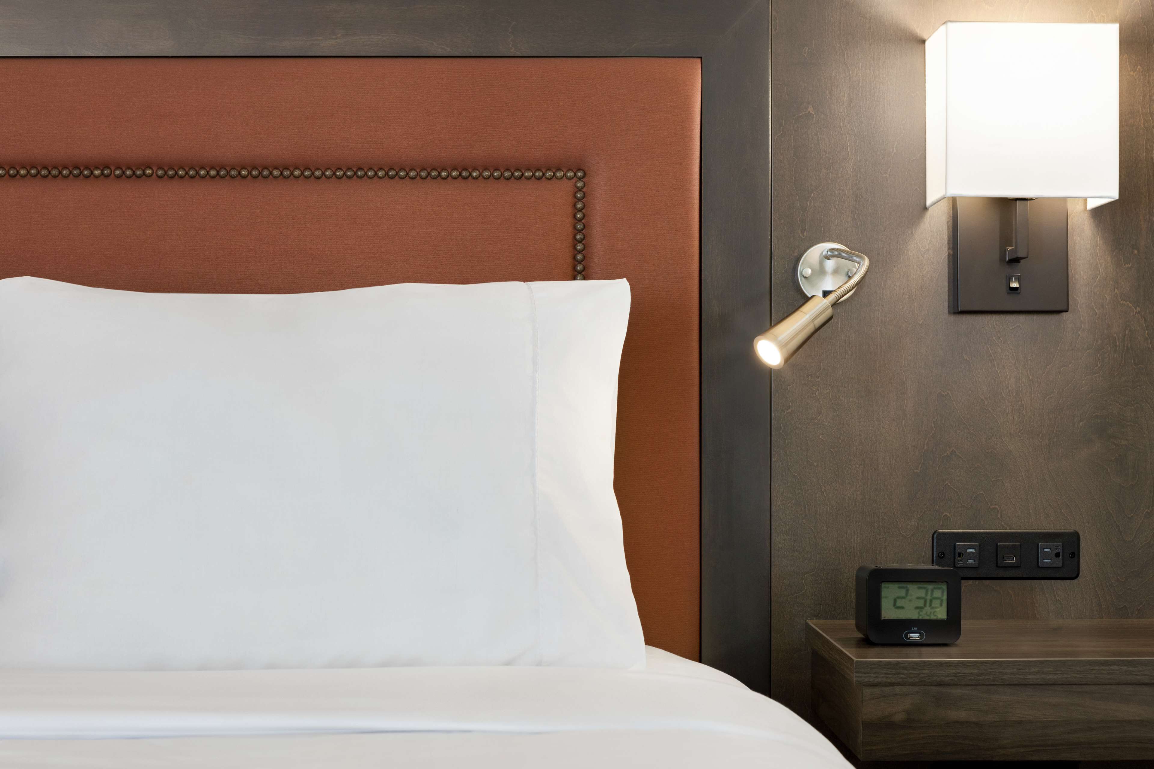 A hotel room with a bed, a nightstand with a digital clock, a wall-mounted lamp, and a wall socket.