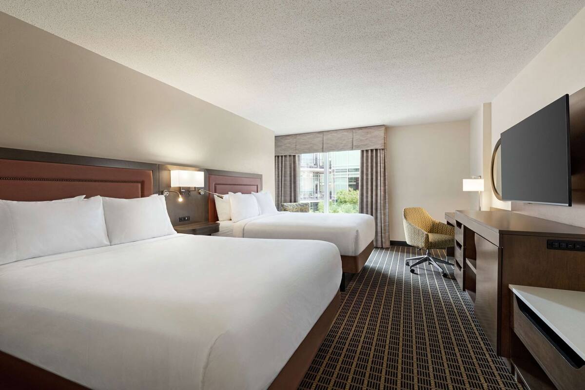 A hotel room with two beds, a desk, a chair, a TV, and a window with curtains.
