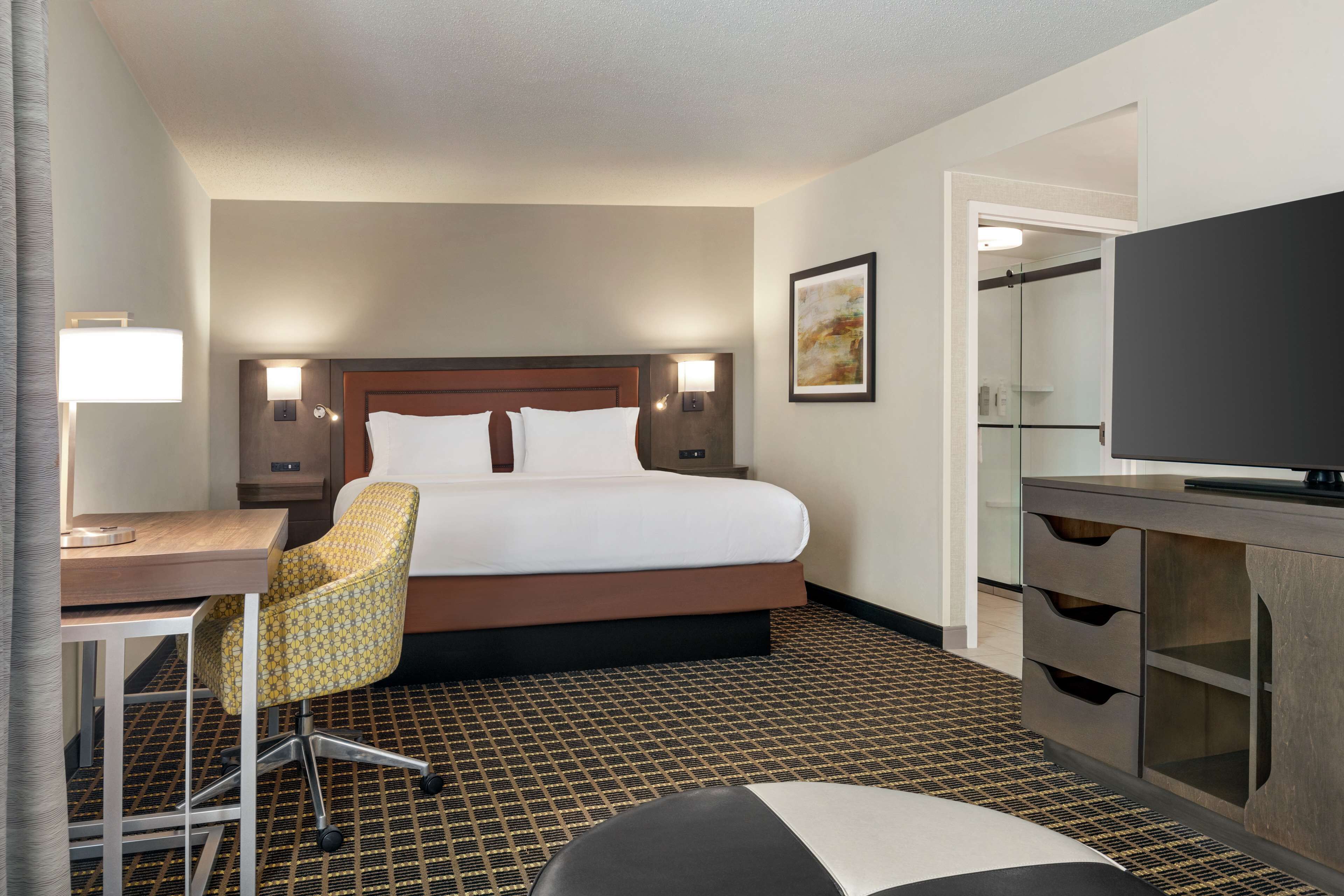 A hotel room with a bed, desk, chair, TV, and a patterned floor.
