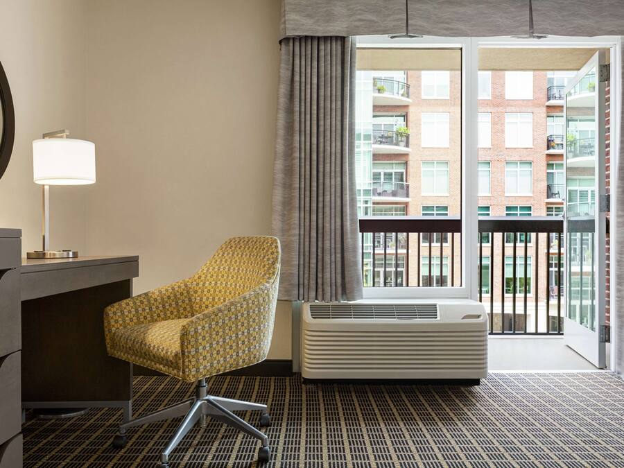 A hotel room with a patterned armchair, a desk with drawers, a standing lamp, an air conditioning unit, and a view of a building through a large window with curtains.
