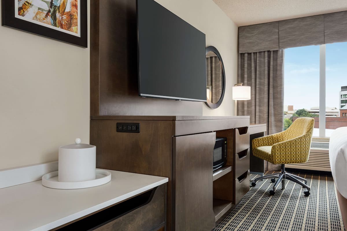 Ein Hotelzimmer mit Flachbildfernseher über einem Holztisch, einer Mikrowelle, einem Sessel und einem großen Fenster mit Blick auf die Stadt.