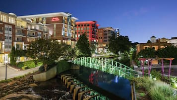 Un hôtel à étages, un pont végétalisé enjambant un bassin d’eau, et un espace extérieur bien éclairé.