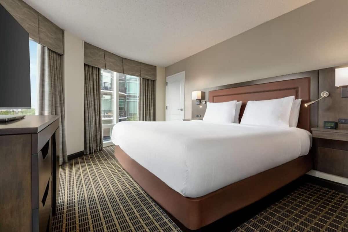 Ein Hotelzimmer mit einem großen Bett, einem Schreibtisch, einem Stuhl, einem Fernseher, einem Fenster mit Vorhängen und einer Wandlampe.