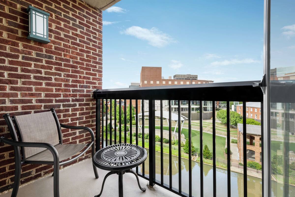 Ein Balkon mit Backsteinmauer, einem Metallstuhl, einem kleinen Tisch und Blick auf ein Gebäude sowie Grünflächen.
