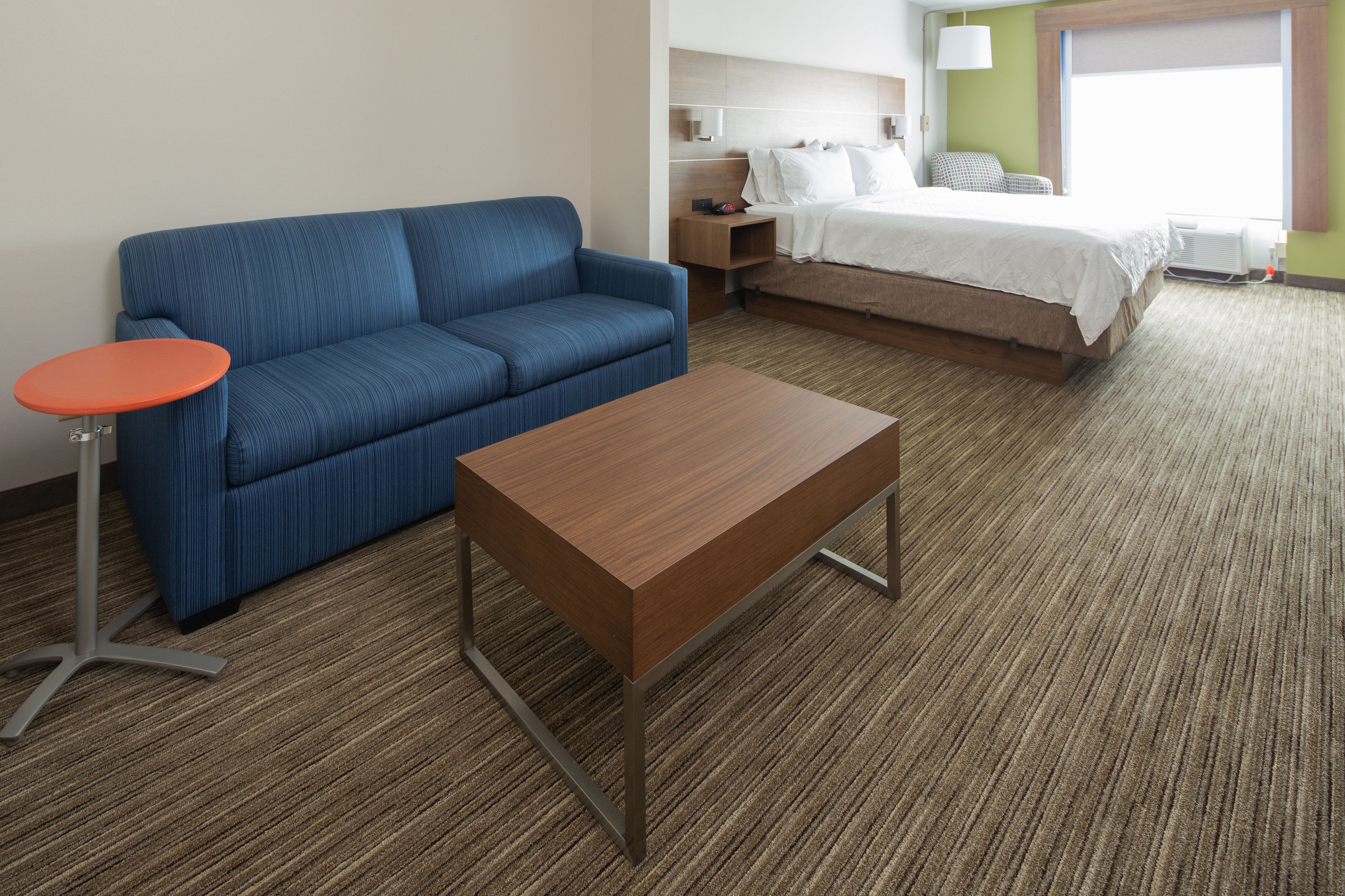 Una habitación de hotel con un sofá azul, una mesita naranja, una mesa de centro de madera, una cama con ropa blanca, una mesita de noche y una lámpara.