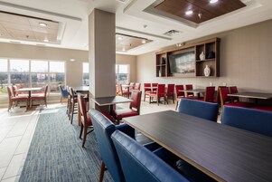 Un comedor de hotel moderno con varias mesas y sillas, amplias ventanas que ofrecen vistas al exterior, y un televisor de pantalla plana montado en la pared.