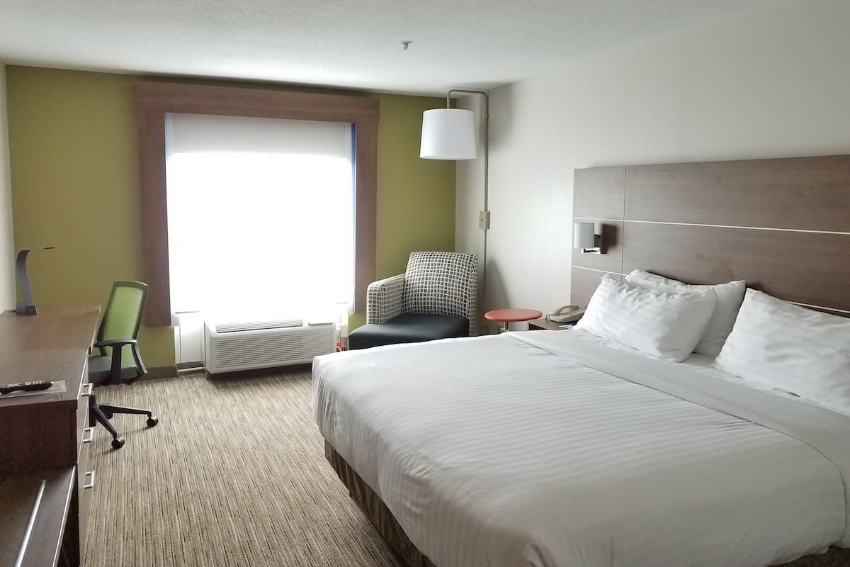 A hotel room with a large bed, a desk with a chair, a small table, and a window with curtains.