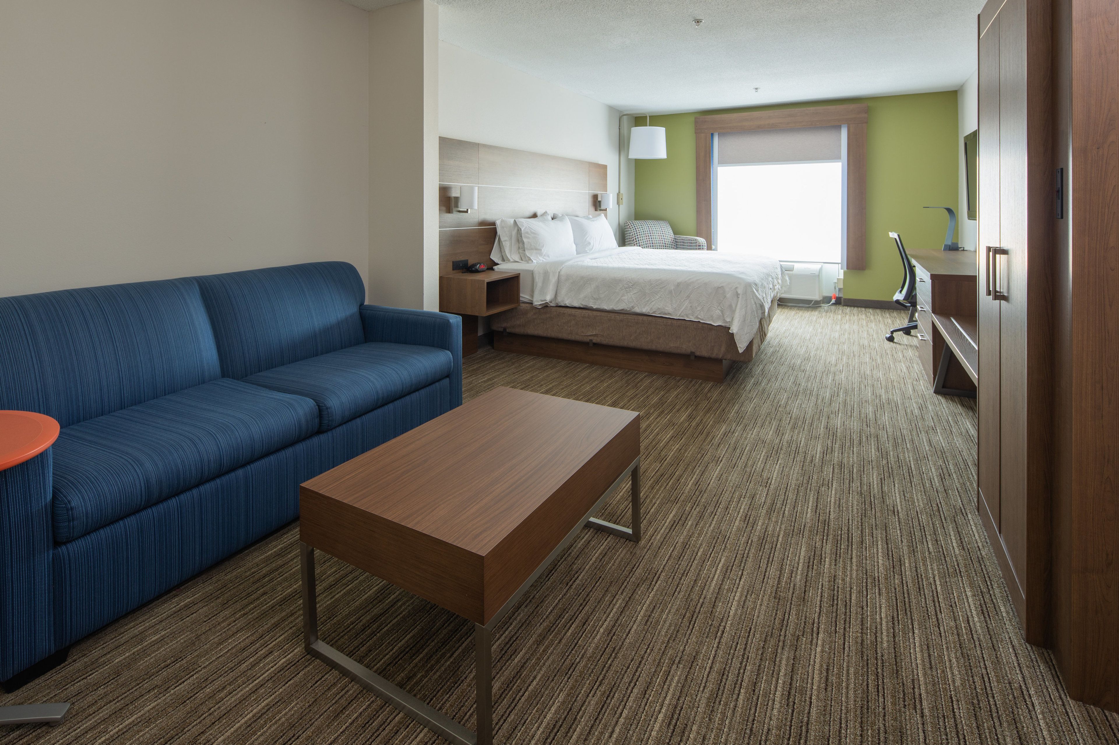 A hotel room with a blue sofa, a wooden table, a bed with white linens, a desk with a lamp, and a view of the outdoors through a window.