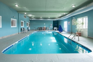 Piscina interior com água azul cristalina, rodeada por paredes pintadas em tom azul claro. Várias janelas permitem a entrada de luz natural.