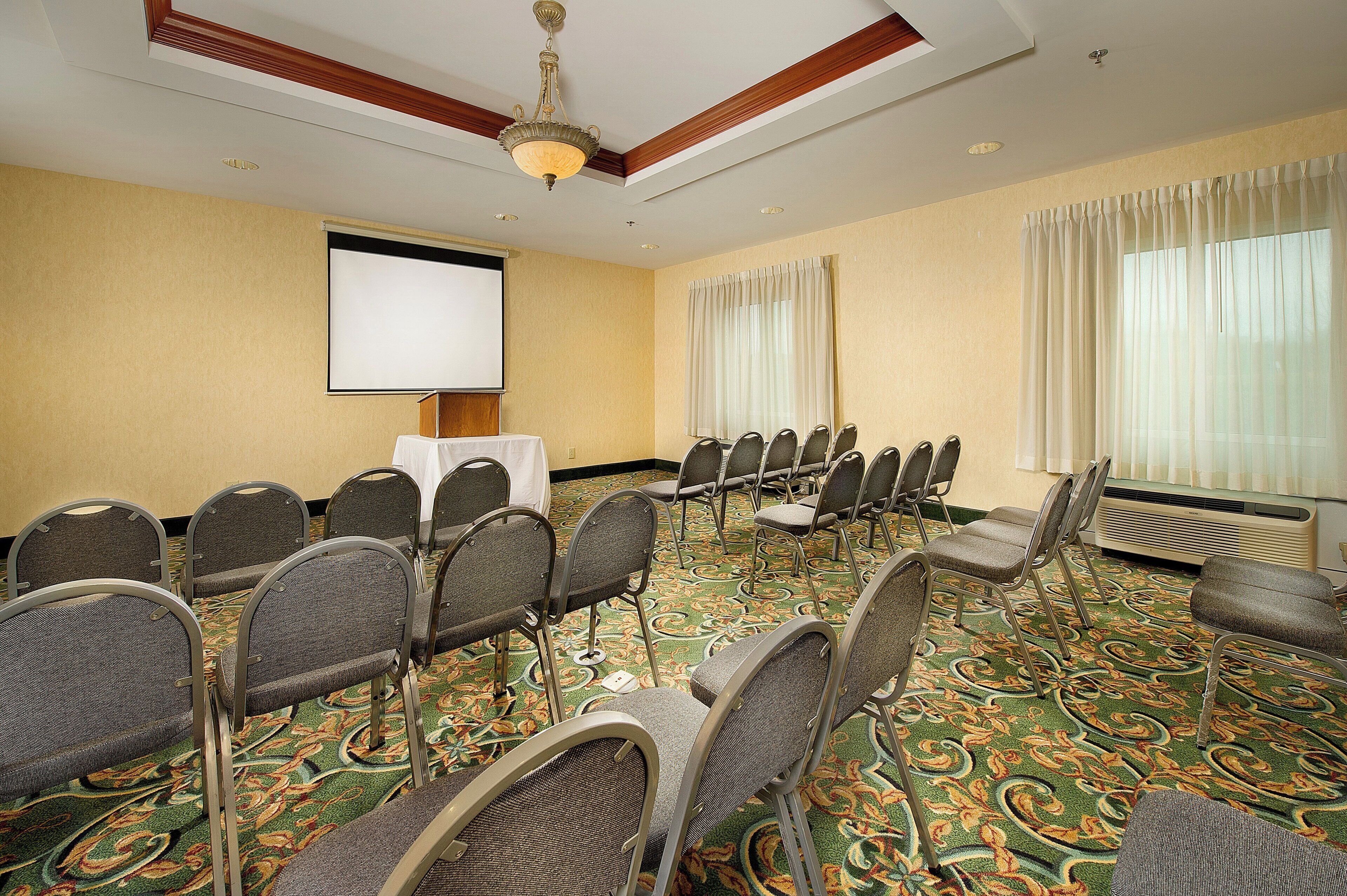 A conference room with a projector screen, rows of chairs, a carpeted floor, and a ceiling fan.