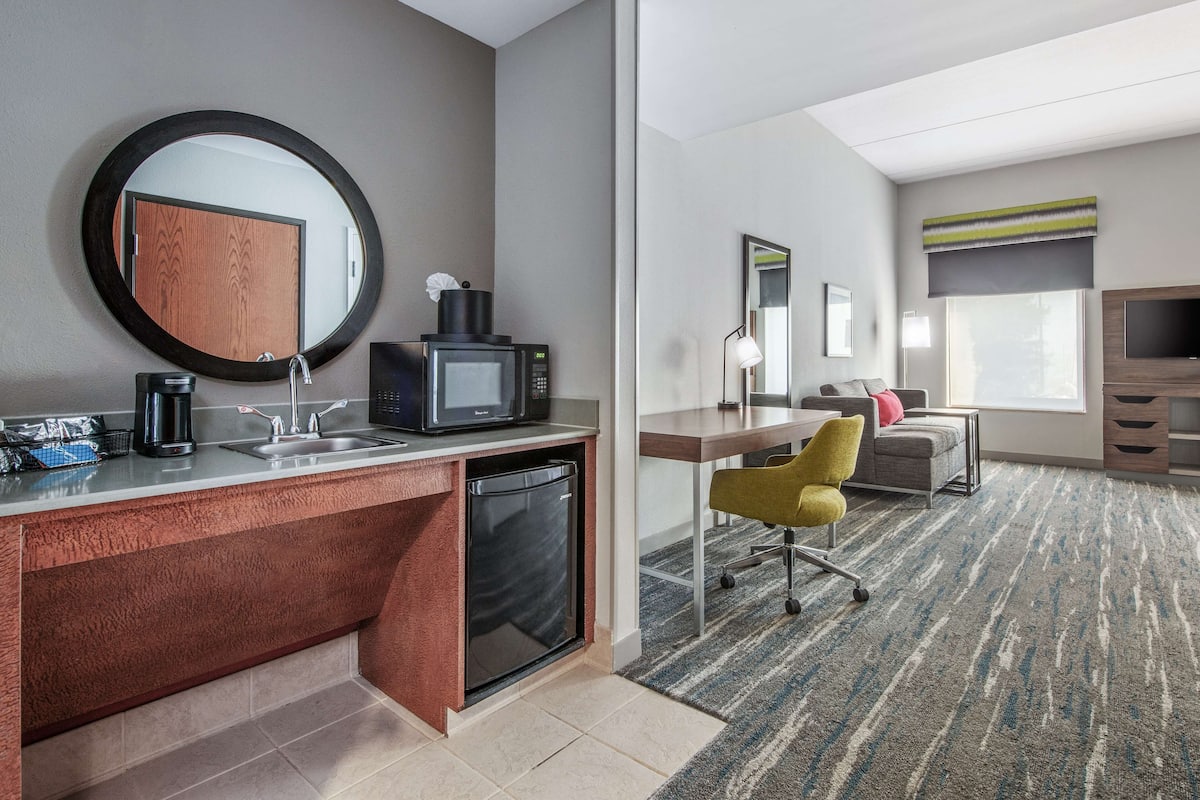 A hotel room with a kitchenette, a desk, a chair, a sofa, a TV, and a large window.