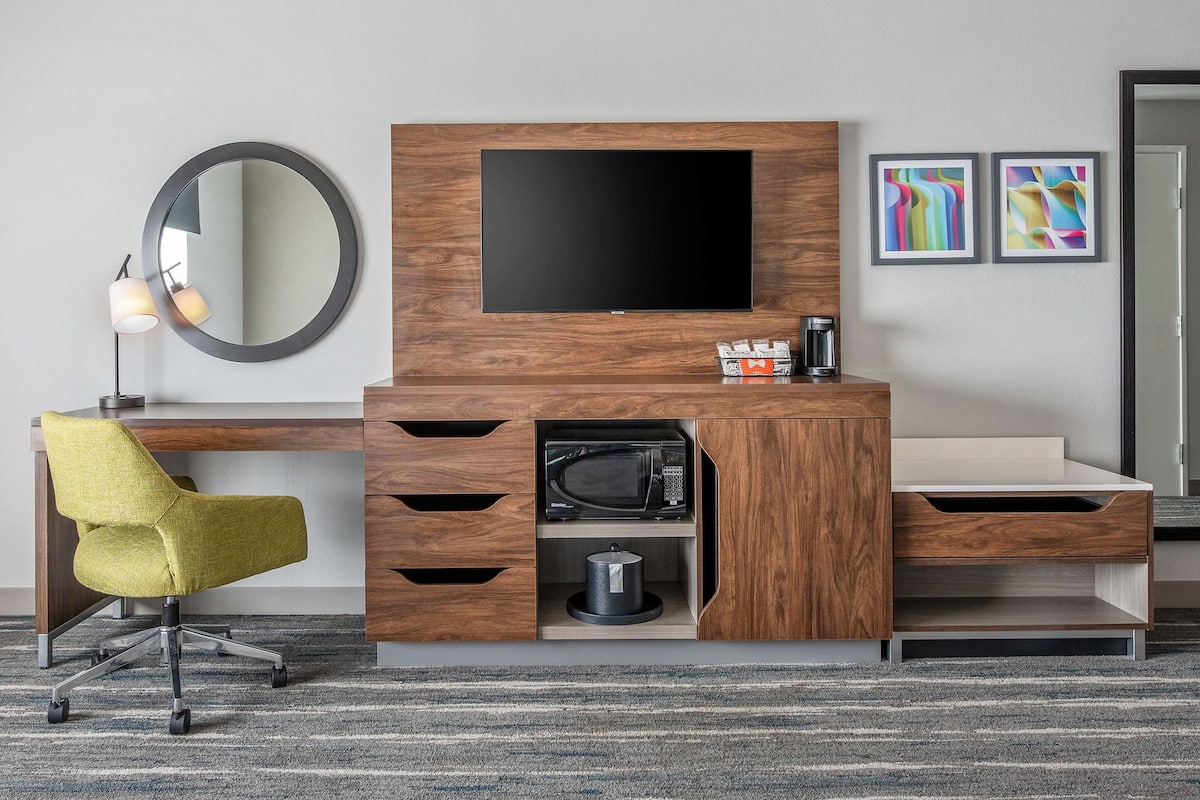 A modern hotel room with a wooden entertainment unit, a flat-screen TV, a microwave, a coffee maker, and a yellow chair.