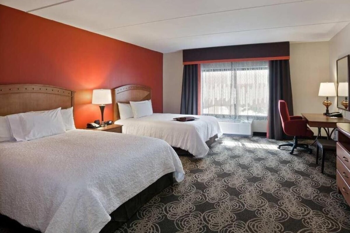 A hotel room with two beds, a desk, a chair, a window with curtains, and a patterned carpet.