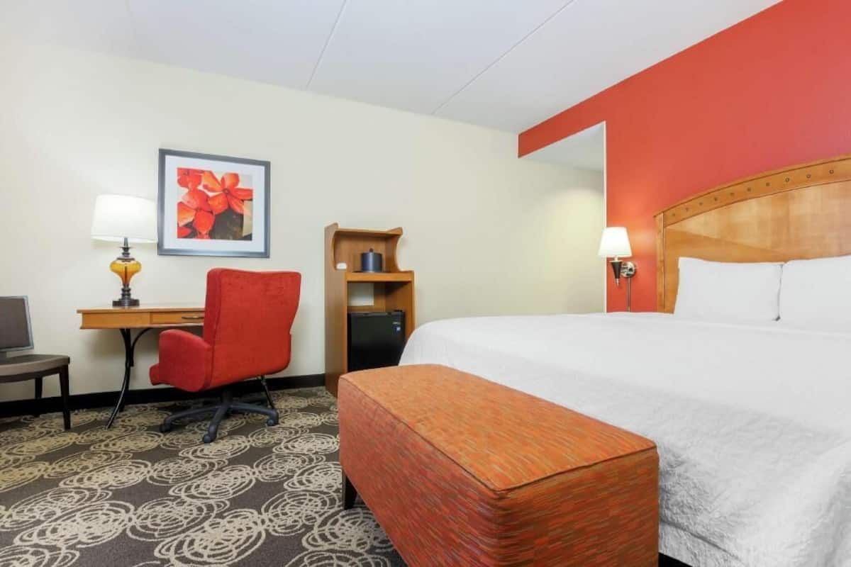 A hotel room with a bed, a desk with a chair, a small cabinet, and a wall-mounted lamp.