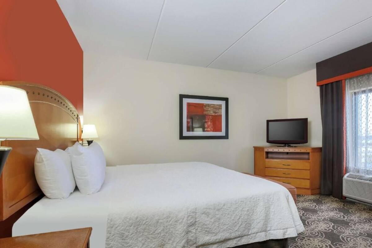 A hotel room with a bed, a television, a framed picture on the wall, and a window with curtains.
