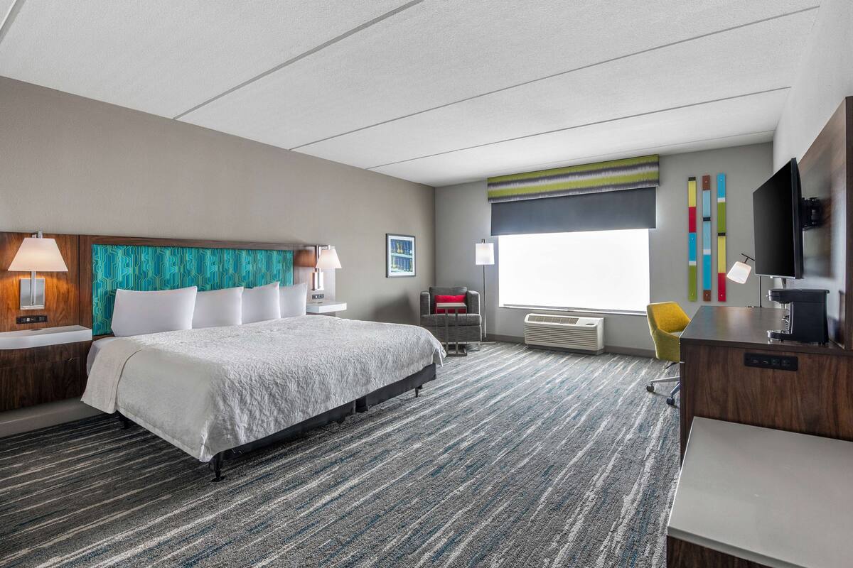 A hotel room with a large bed, a desk, a chair, a TV, and a window with blinds.