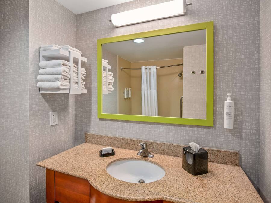 A bathroom with a granite countertop sink, a green-framed mirror, and neatly folded towels on a shelf.