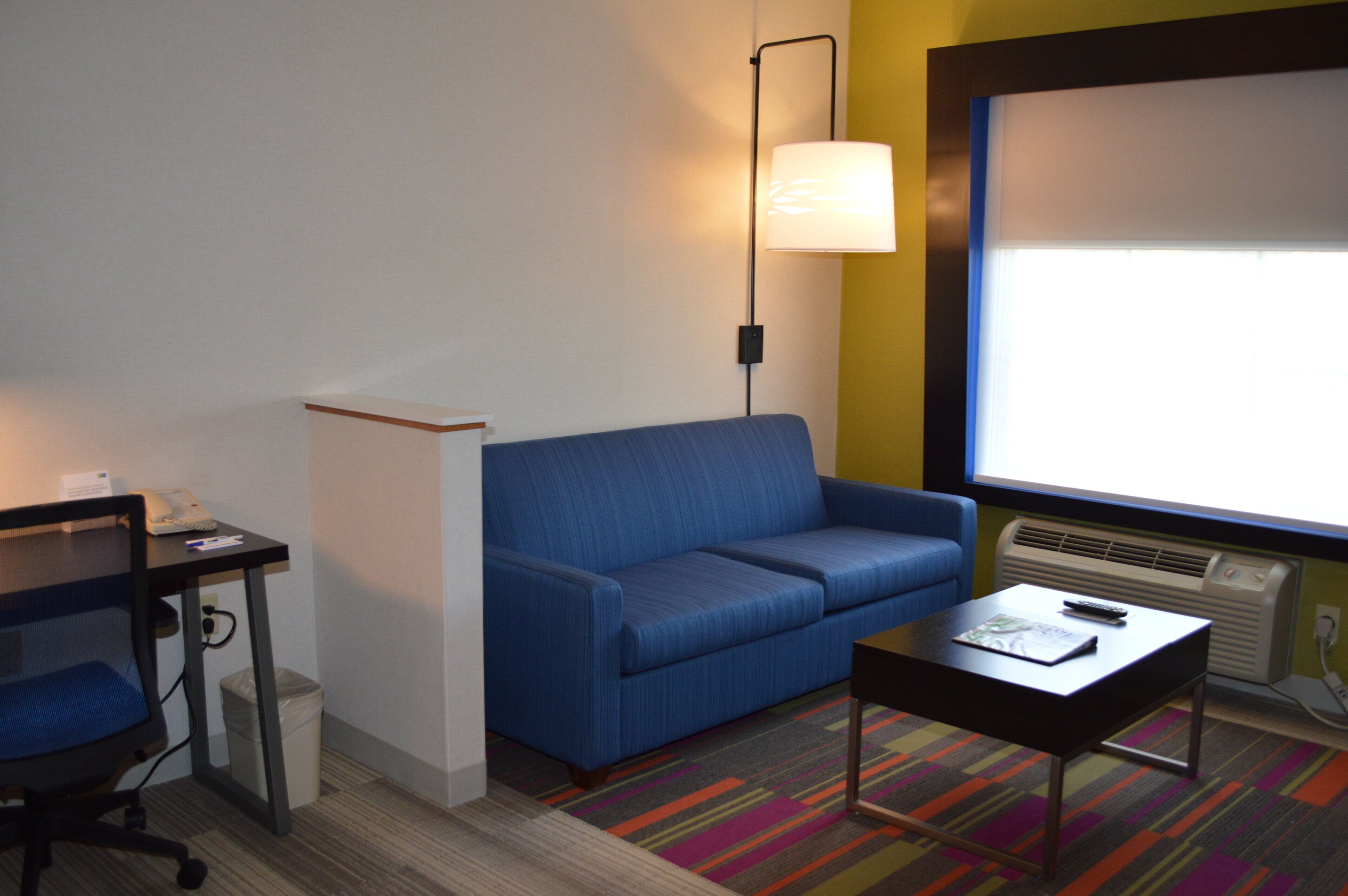 A hotel room with a blue sofa, a small table, a desk with a phone, a lamp, and a projector screen.