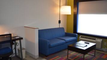 A hotel room with a blue sofa, a small table, a desk with a phone, a lamp, and a projector screen.