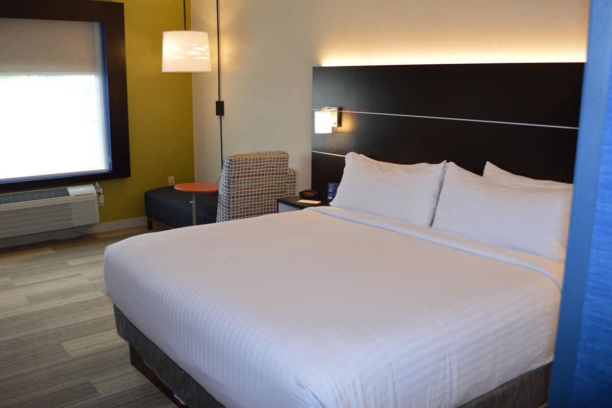 A hotel room with a large bed, a nightstand, a chair, a lamp, and a window with blinds.
